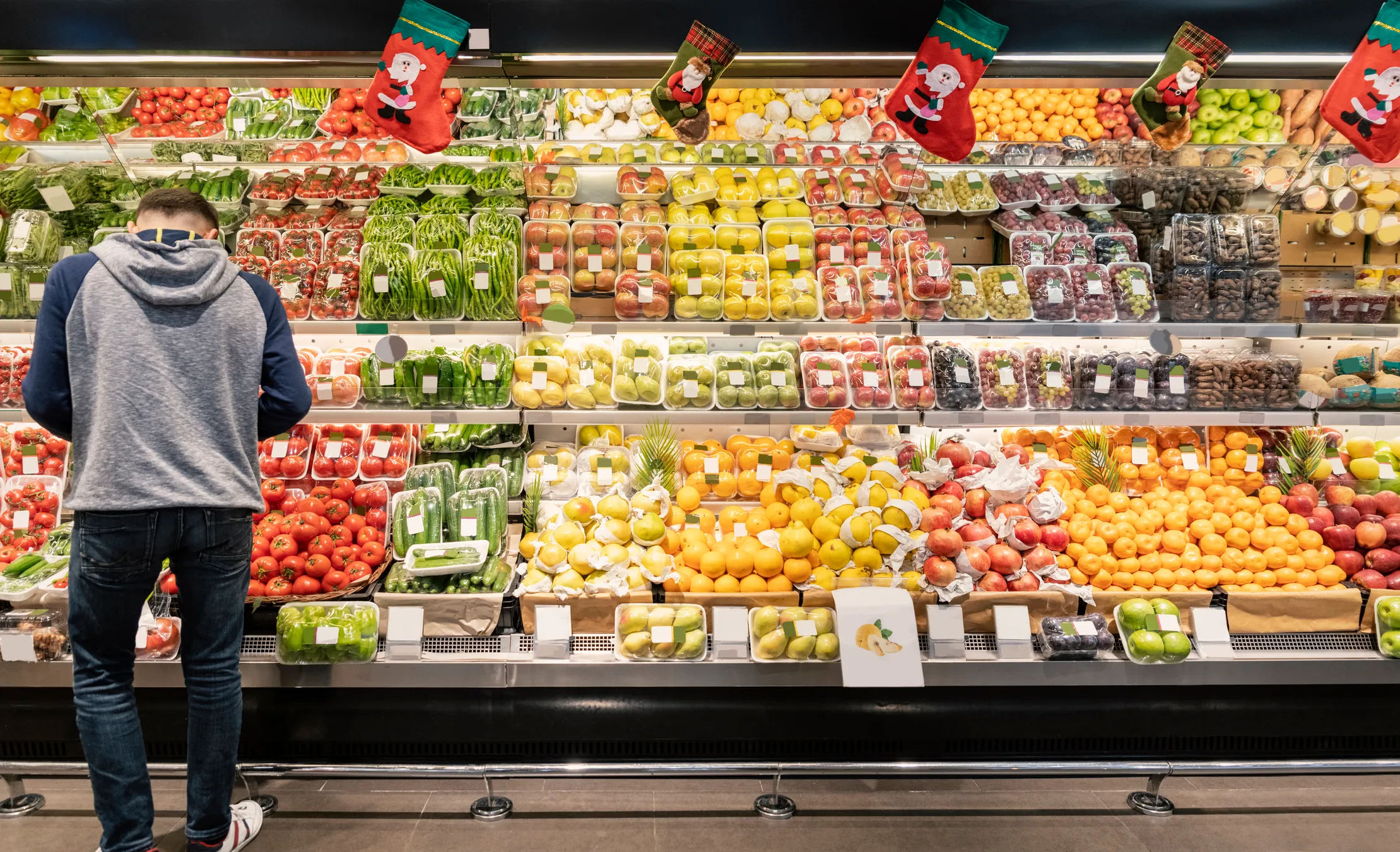 Man wearing a hoodie and jeans standing in front of a grocery store produce display filled with packaged fruits and vegetables including tomatoes, green beans, apples, citrus fruits, and grapes, with Christmas stockings hanging above.