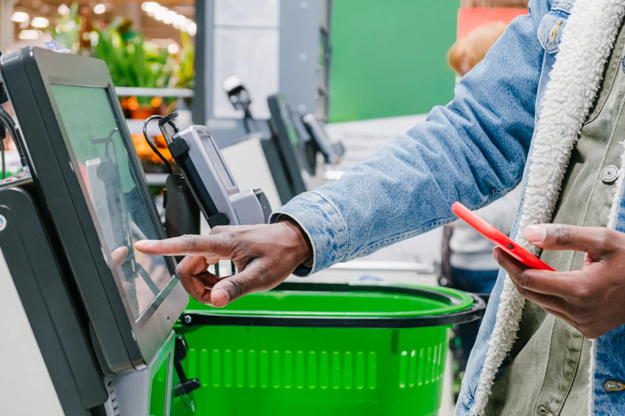 Person using a self-checkout touchscreen at a grocery store while holding a red smartphone, with a green shopping basket in the background.