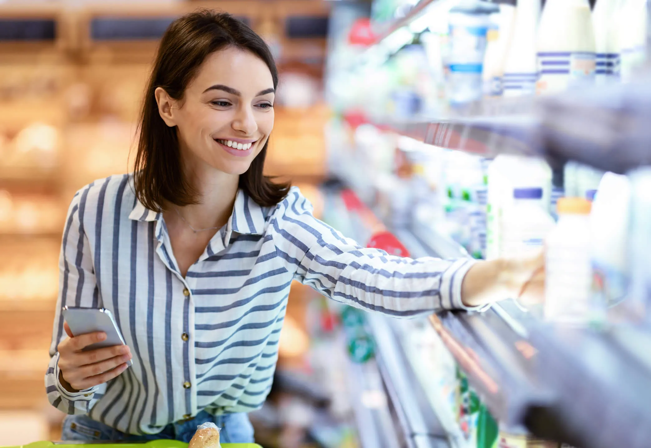 Smiling woman in a striped shirt holding a smartphone while reaching for an item on a refrigerated grocery store shelf.