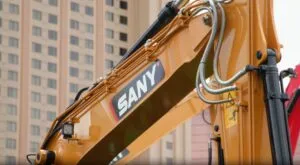 Close-up of a yellow SANY excavator arm with hydraulic tubes and a blurred building in the background.
