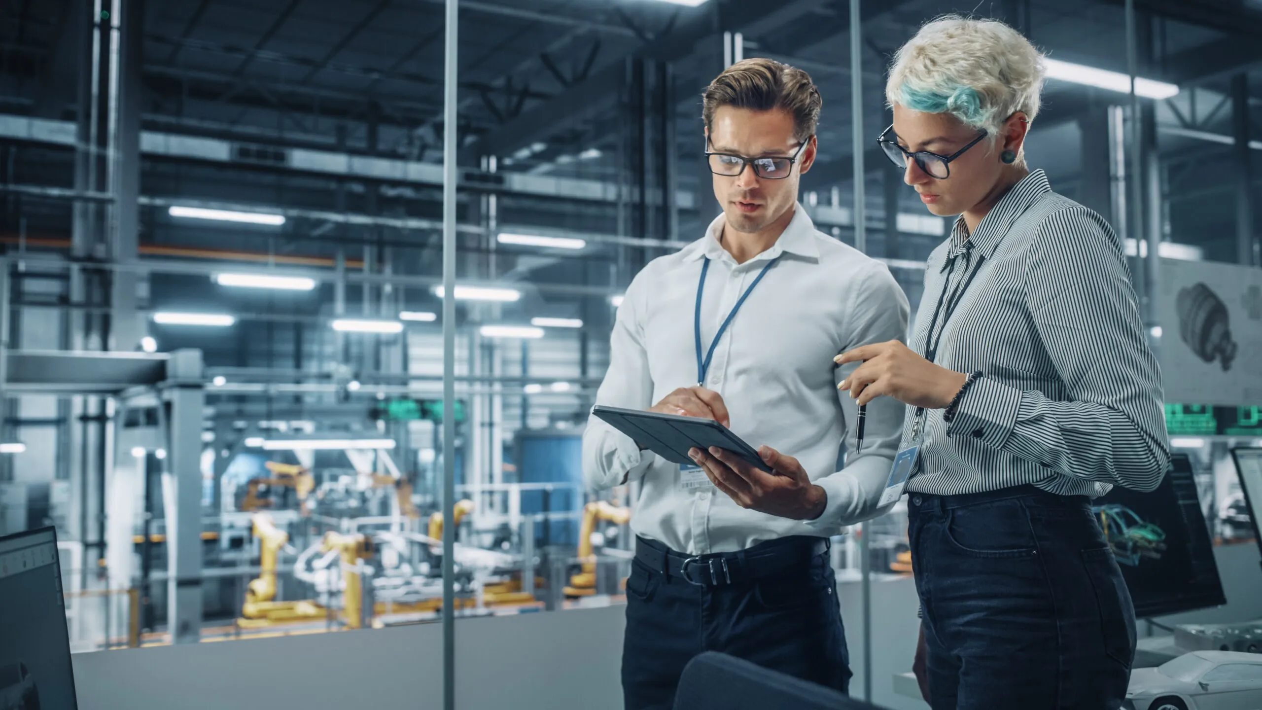 Two engineers wearing glasses and ID badges review a tablet in a modern industrial facility with robotic arms visible through glass behind them.