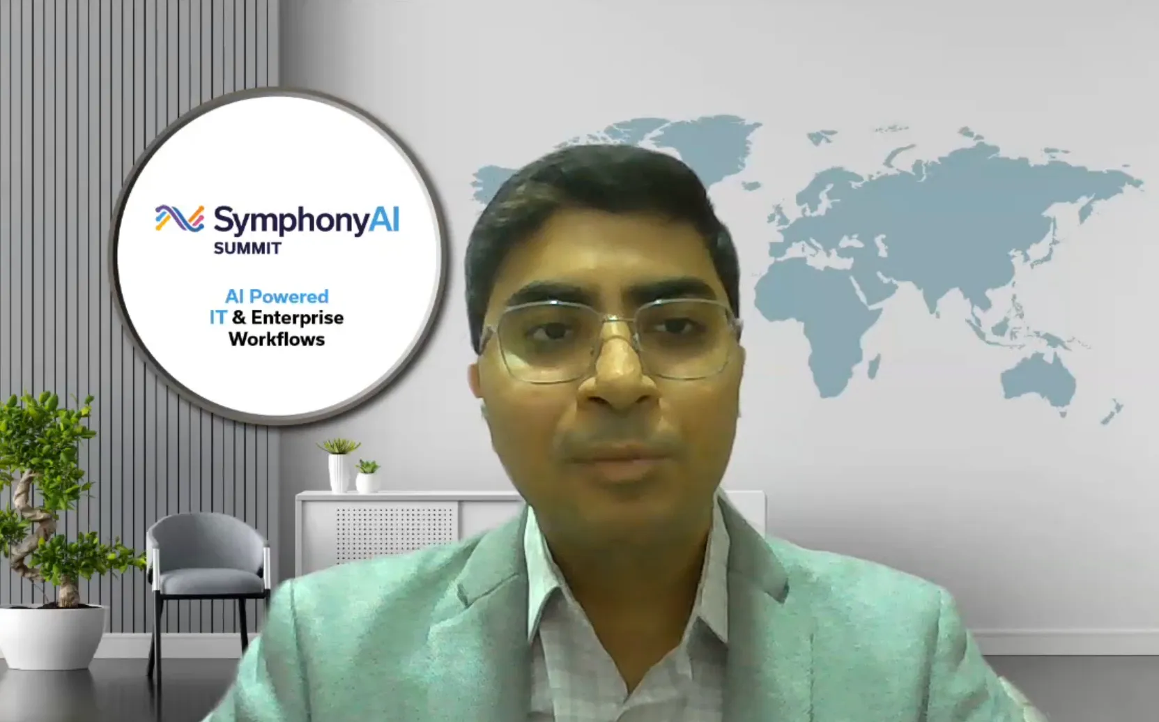 Man wearing glasses and a light gray blazer speaking in front of a virtual background with a map of the world and a round sign reading 'SymphonyAI Summit AI Powered IT & Enterprise Workflows'.