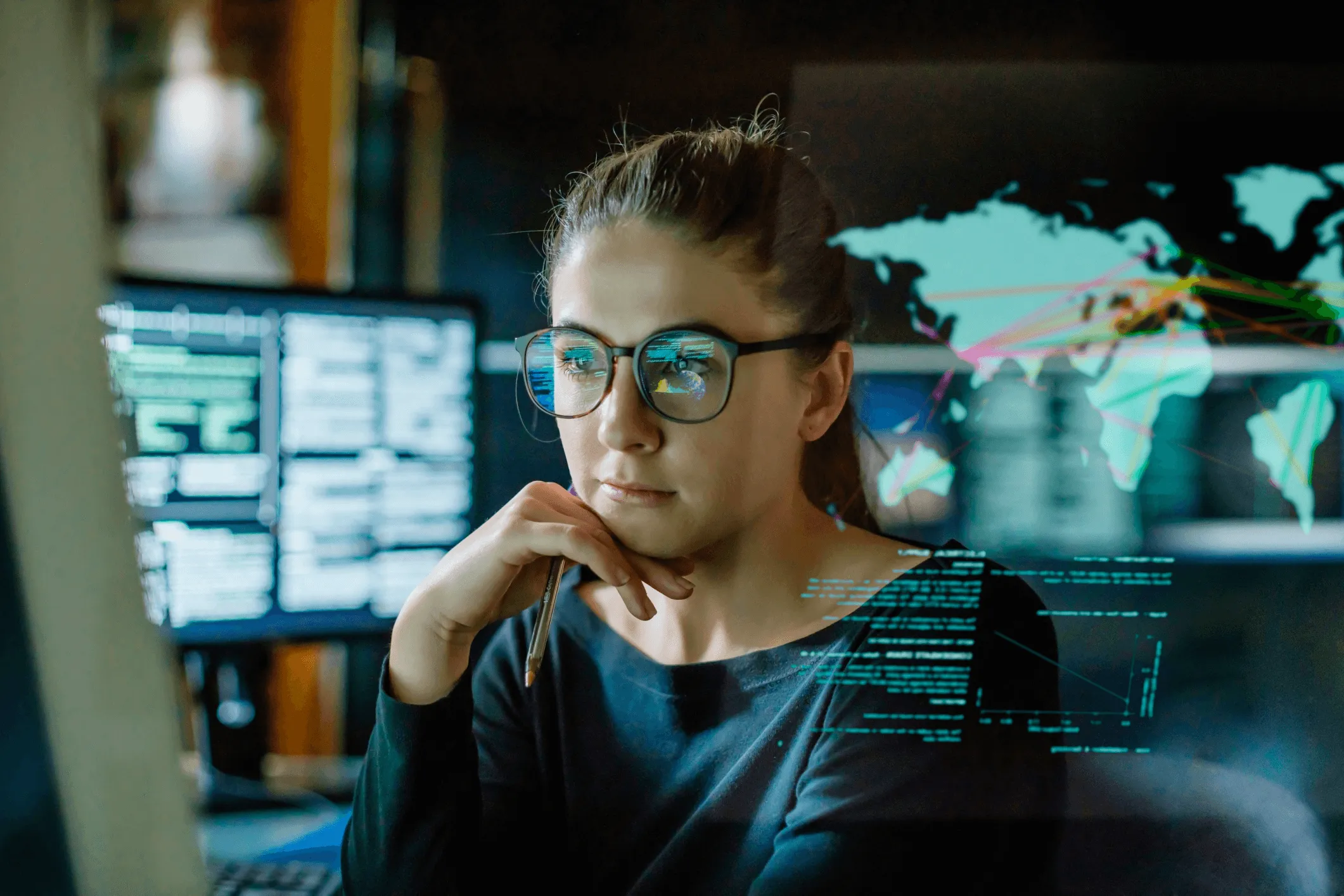 Woman with glasses holding a pen, looking thoughtfully at computer screens displaying code and a world map with network connections.