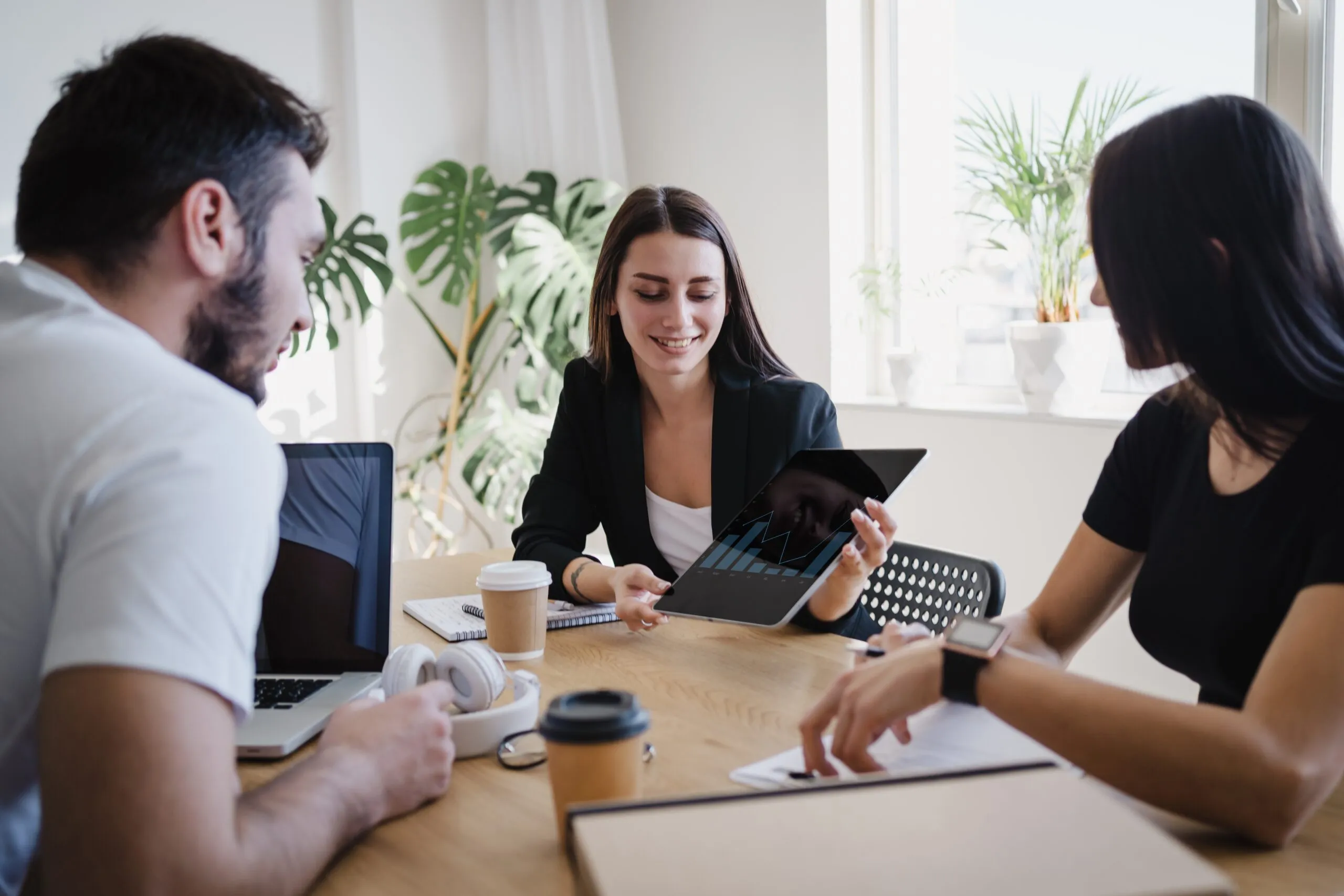 Three young professionals sitting around a table with laptops and coffee, a woman holds a tablet displaying a bar and line graph while smiling, in a bright office with large windows and plants.