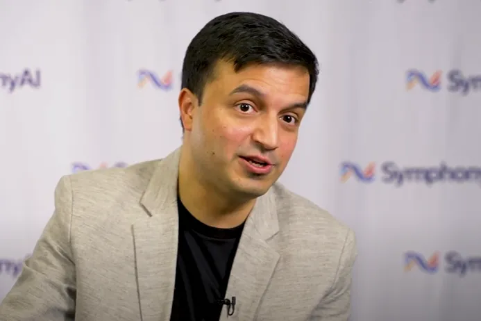 A man with short black hair wearing a light gray blazer and black shirt speaking in front of a backdrop with the SymphonyAI logo repeated on it.