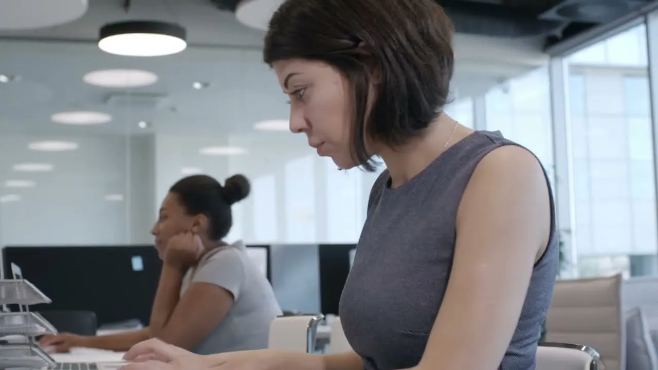 Two women working at desks in a modern office with large windows and overhead lights, both focused on their computers.
