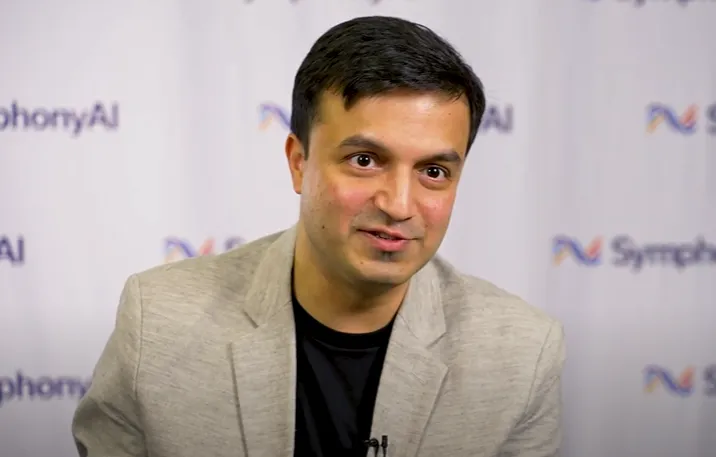 Man with short dark hair wearing a beige blazer and black shirt speaking in front of a backdrop with blurred SymphonyAI logos.