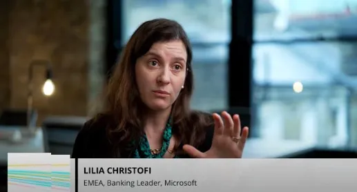 A woman with long brown hair wearing a black top is speaking and gesturing with her right hand in an indoor office setting with large windows behind her. Text overlay identifies her as Lilia Christofi, EMEA Banking Leader at Microsoft.