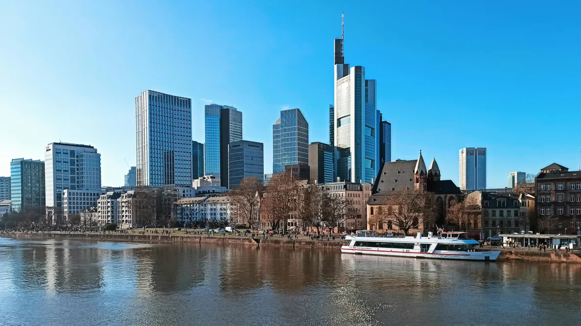 View of Frankfurt skyline with modern skyscrapers and historic buildings along a riverbank under a clear blue sky, including a docked white riverboat.