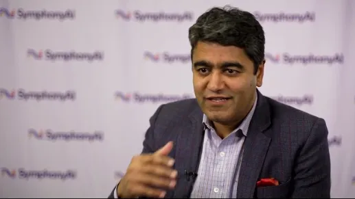 A man with black hair wearing a dark checkered blazer and light-colored shirt speaks in front of a backdrop featuring the SymphonyAI logo repeated several times.
