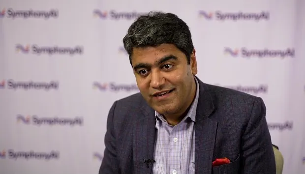 A man with gray and black hair, wearing a plaid jacket and checked shirt, sitting and talking in front of a backdrop with repeated SymphonyAI logos.