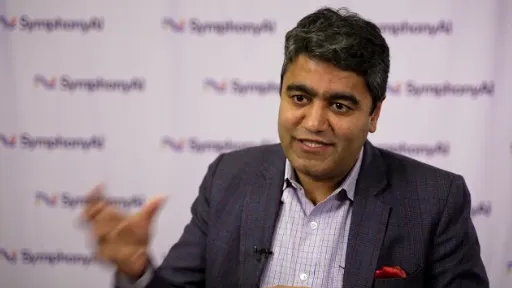 A man with dark hair in a checkered blazer and light shirt gestures with his right hand while speaking, with a blurred background featuring repeated SymphonyAI logos.