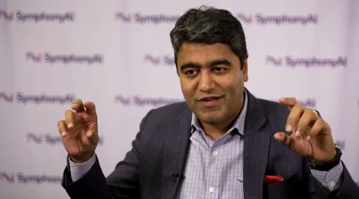 A man with dark hair and wearing a dark blazer and checked shirt is speaking with his hands raised, making a gesture with both hands, seated in front of a backdrop with repeated SymphonyAI logos.