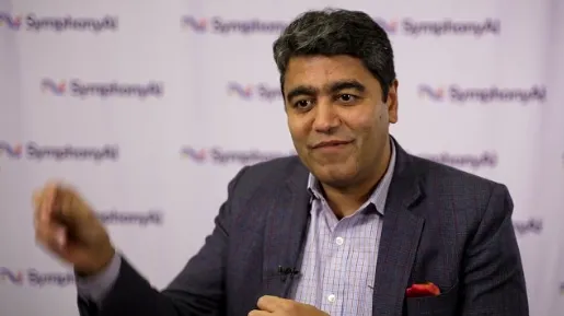 Man with short dark hair and dark blazer speaking in front of a white backdrop with repeated SymphonyAI logos.