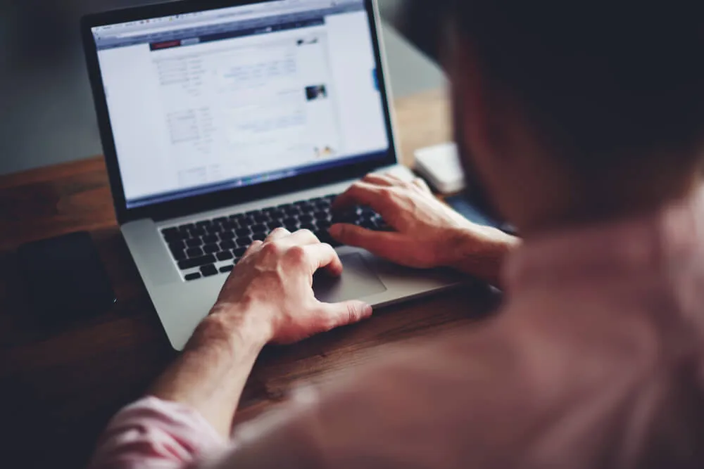 Person typing on a laptop with a webpage open displaying text and small images, viewed from behind over the shoulder.