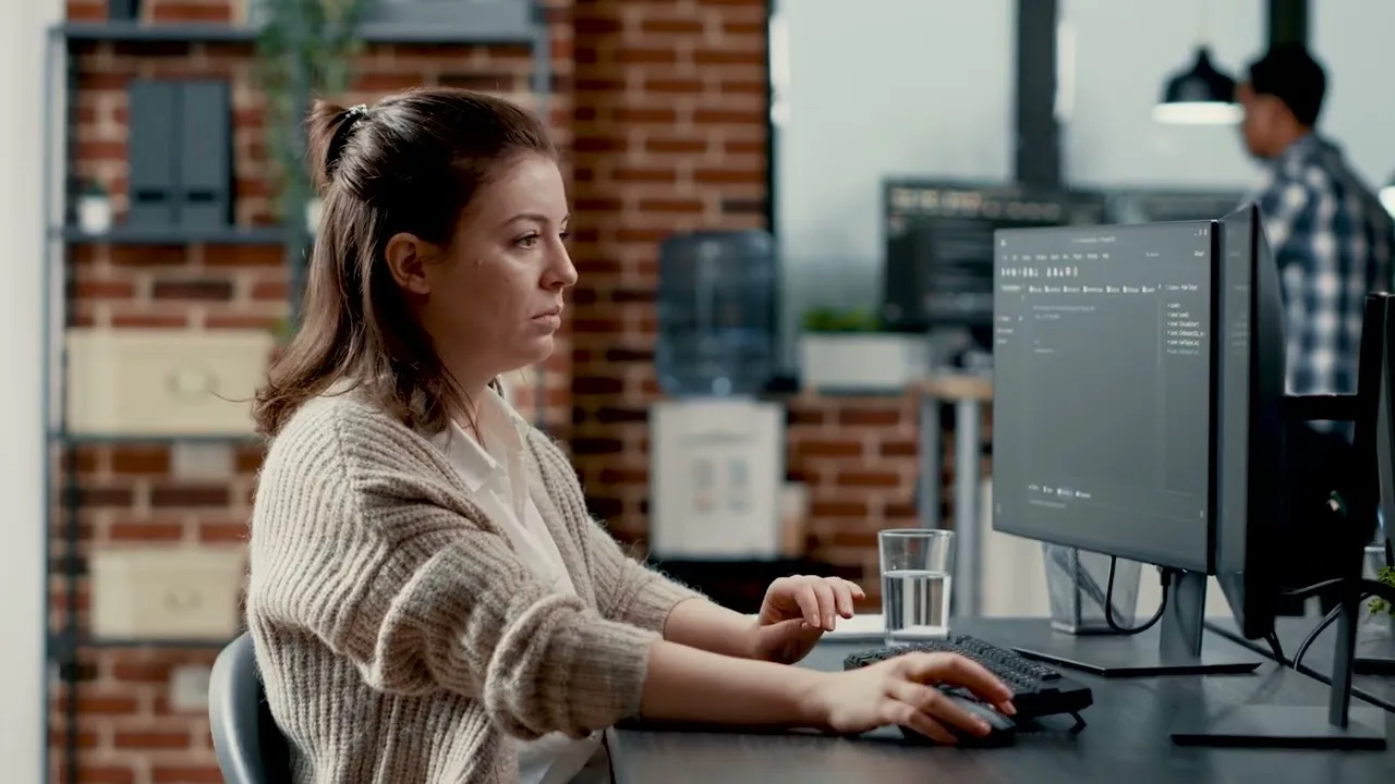 Woman in a beige sweater working on a desktop computer with multiple monitors in an office with exposed brick walls, a water dispenser, and a colleague in the background.