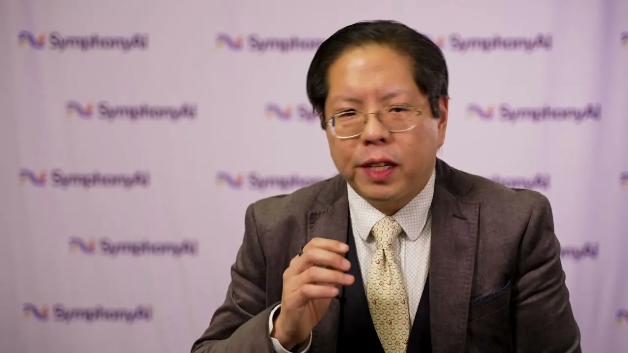 Man with glasses wearing a brown suit, white shirt, and patterned tie, speaking in front of a backdrop with repeated SymphonyAI logos.