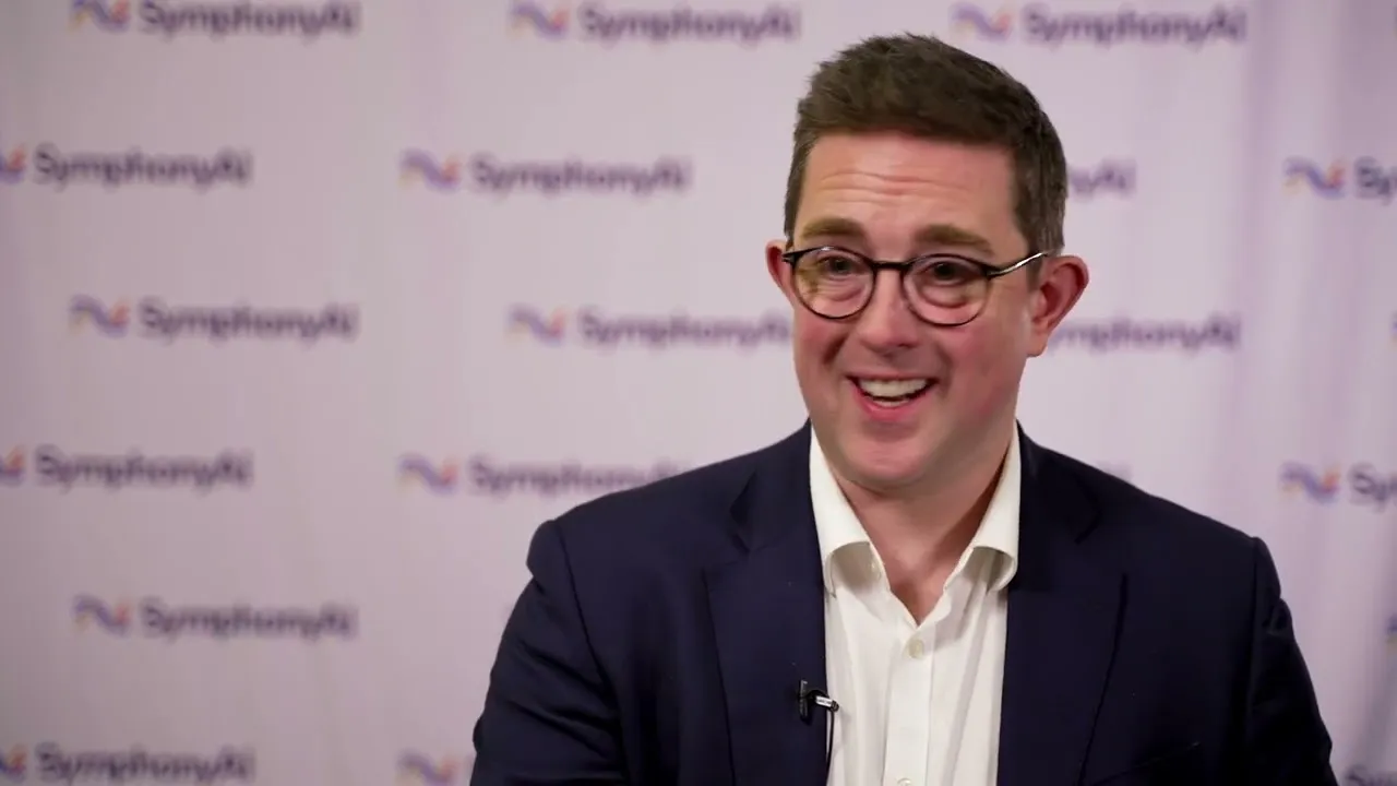 Smiling man with short brown hair and glasses wearing a navy blazer and white dress shirt, sitting in front of a backdrop with repeated SymphonyAI logos.