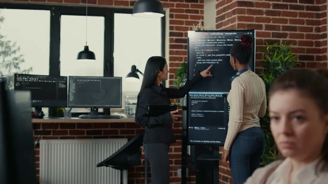 Two women in a modern office are looking at and discussing code displayed on a large vertical monitor mounted on a brick wall, while a third woman is in the foreground slightly blurred.