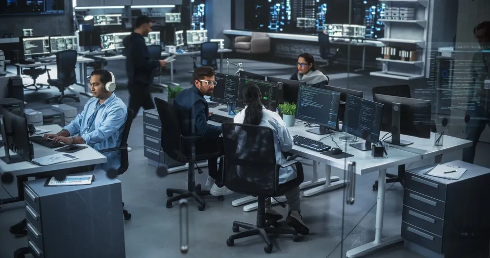 A modern office with multiple workers at computer desks surrounded by monitors displaying code, with a professional atmosphere of collaboration and technology.
