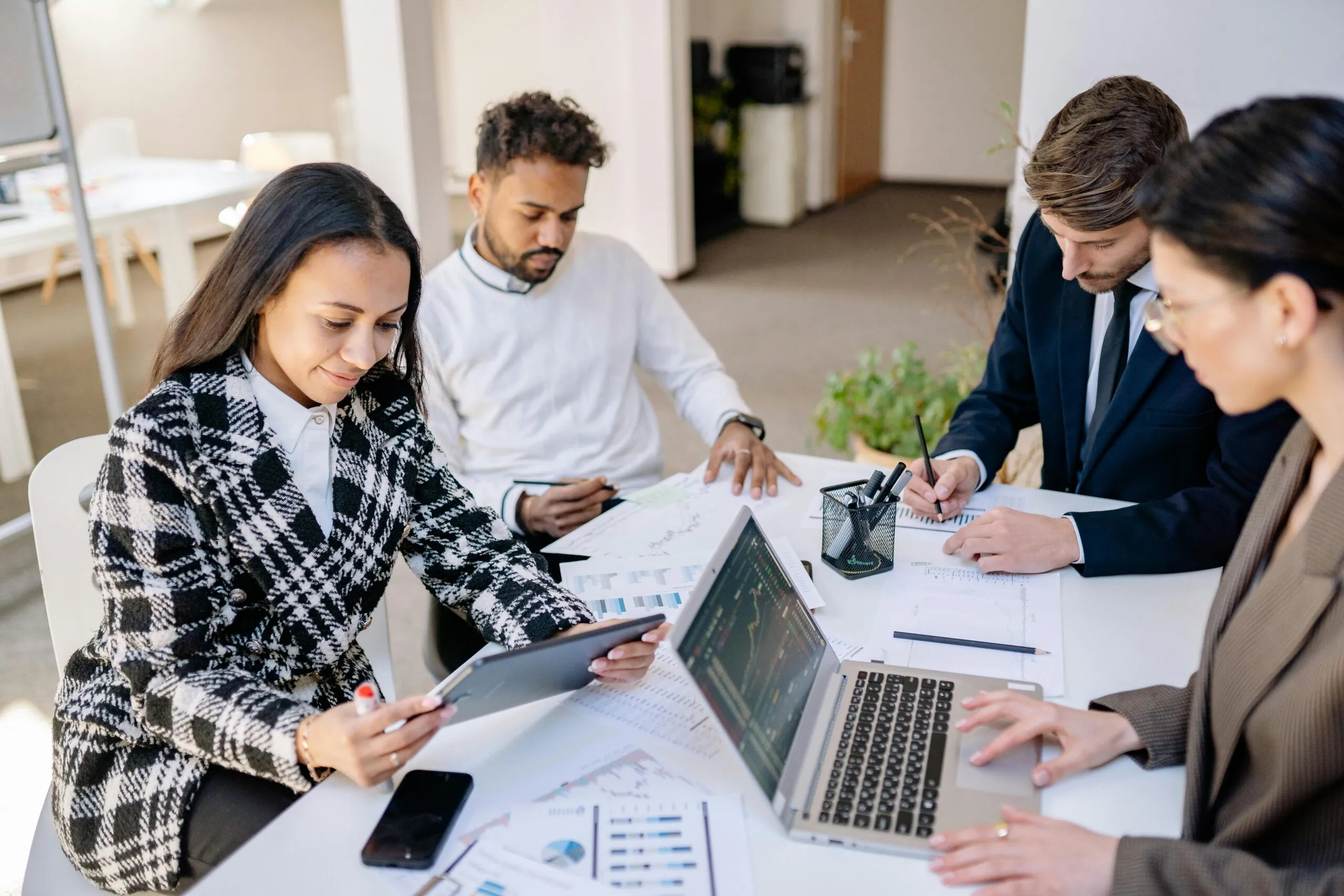 Four diverse business professionals working together at a table with laptops, tablets, charts, and documents, focusing on data analysis and discussions in a modern office.