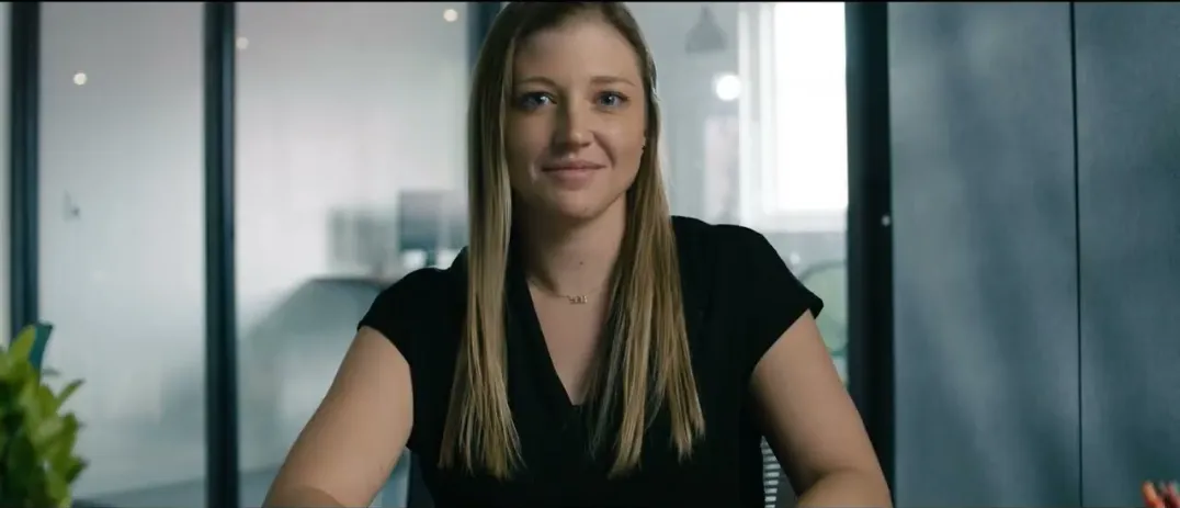 A woman with long blonde hair wearing a black shirt, sitting and facing the camera with a slight smile in an office setting.