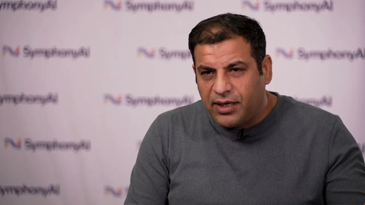 Man in a gray long-sleeve shirt speaking in front of a blurred background with repeated SymphonyAI logos.