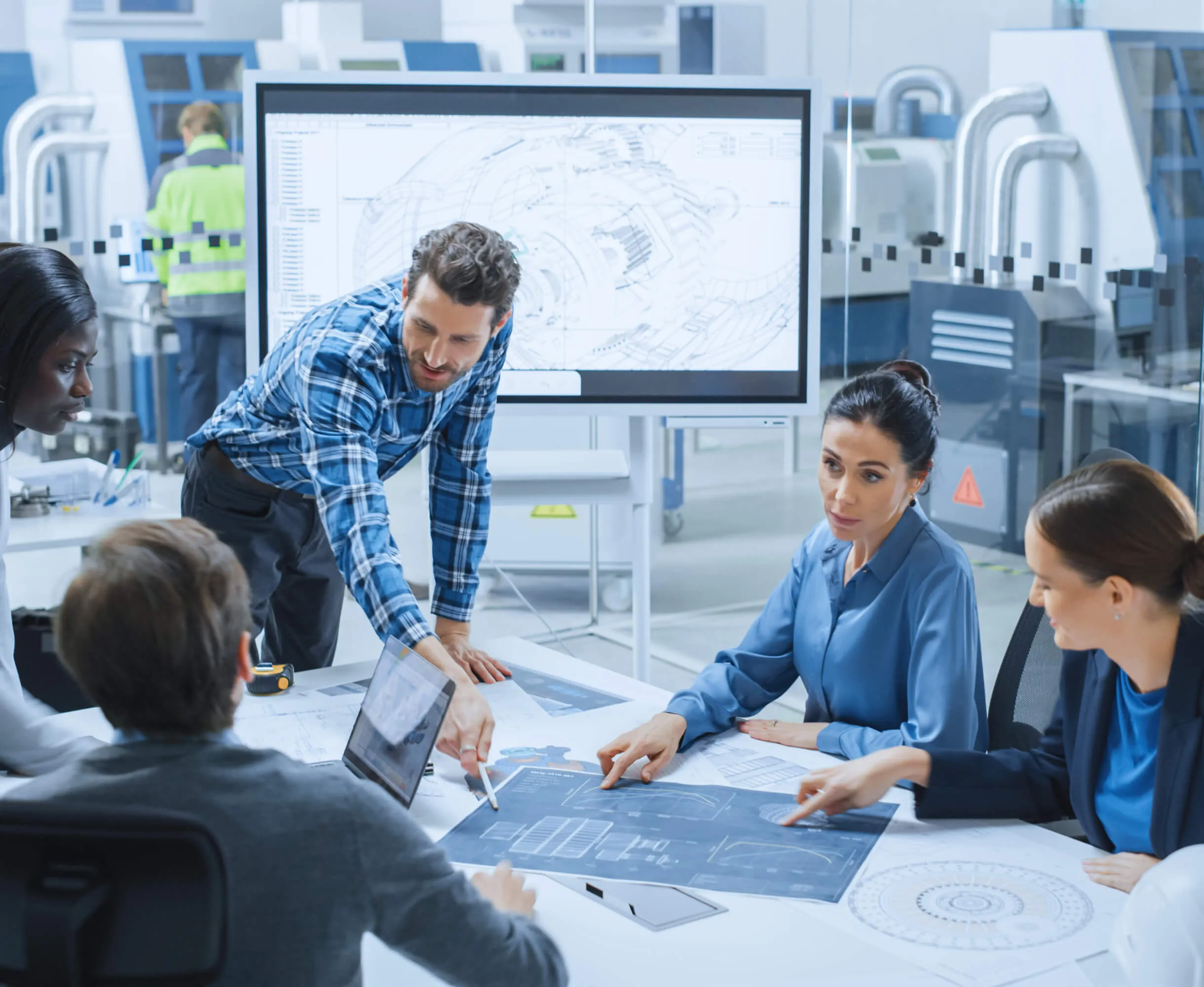 Four engineers gathered around a white table with technical blueprints and a laptop, discussing a project in a modern industrial workspace with machinery and a large monitor displaying a technical drawing in the background.