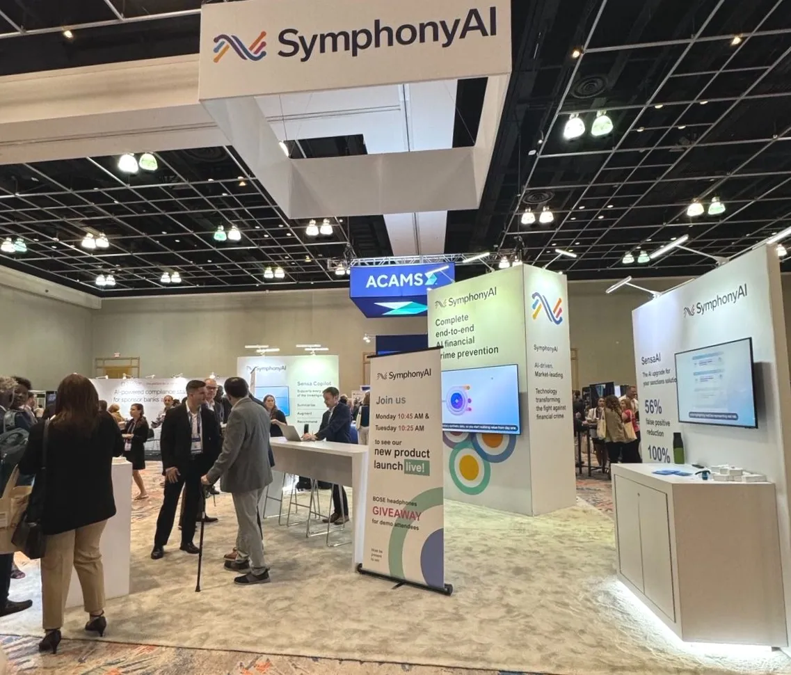 Conference exhibition booth for SymphonyAI with multiple lighted panels displaying company logo and product information, including AI-driven financial crime prevention and SensaAI features, a screen showing graphics, and a banner about a new product launch and Bose headphones giveaway. Several people are engaging and conversing at the booth in a large indoor event space with overhead lighting and an ACAMS sign in the background.