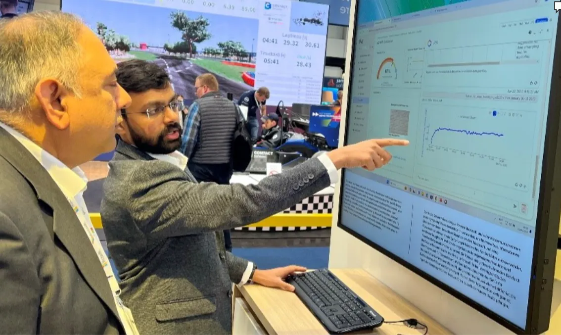 Two men in business attire are looking at a large computer screen, one pointing at data charts and graphs displayed on the screen during a trade show or tech event, with racing simulator screens and people in the background.