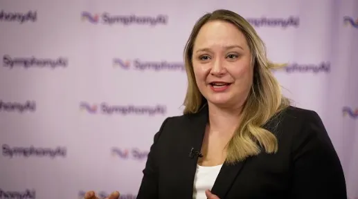 A woman with long blonde hair wearing a black blazer and white shirt, speaking in front of a backdrop with repeated SymphonyAI logos.