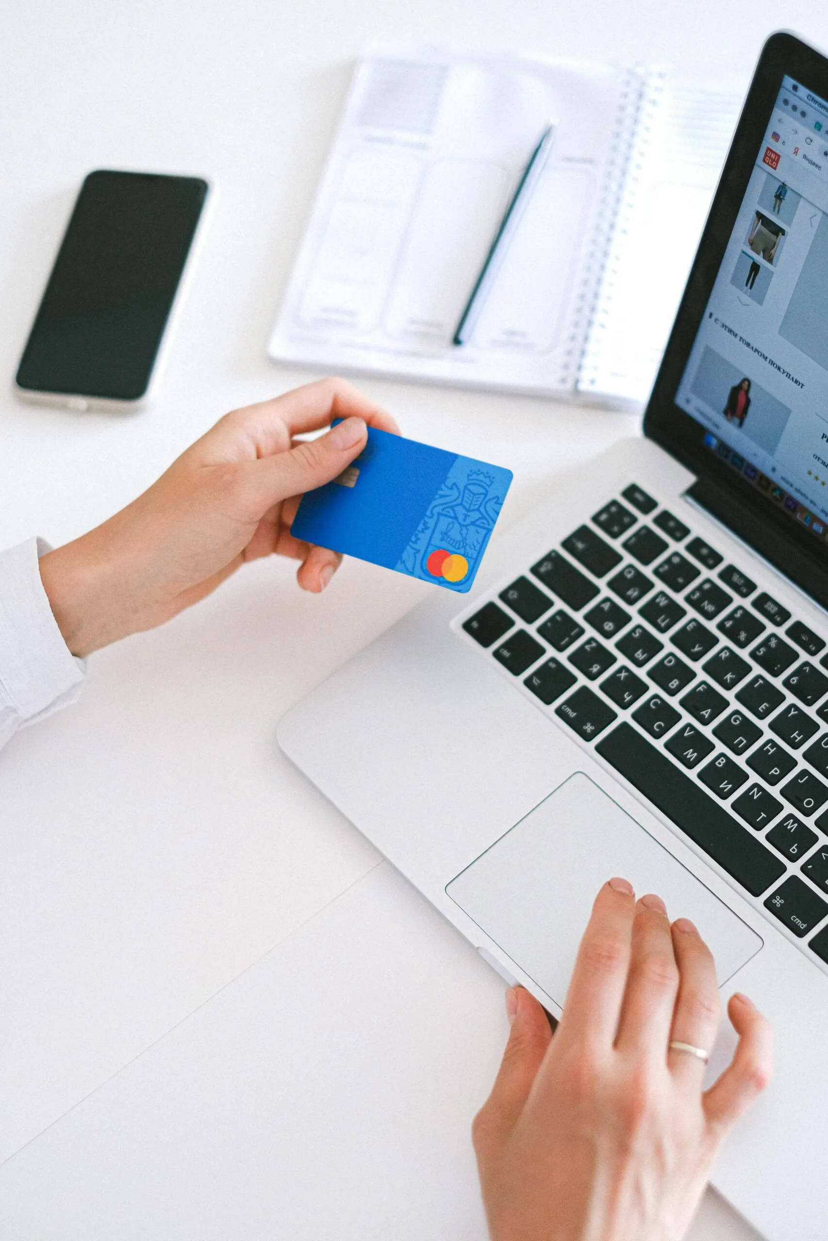 Person holding a blue credit card with a Mastercard logo near a laptop keyboard, with a phone and open notebook with a pen on a white desk.