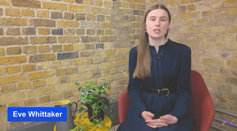 A woman with long light brown hair wearing a dark blue dress with a belt sits on a red chair against a yellowish brick wall, speaking. A yellow table with a green-leaved plant, a yellow mug, and a gray owl-shaped object is next to her. The name 'Eve Whittaker' appears in white text on a blue label in the bottom left corner.