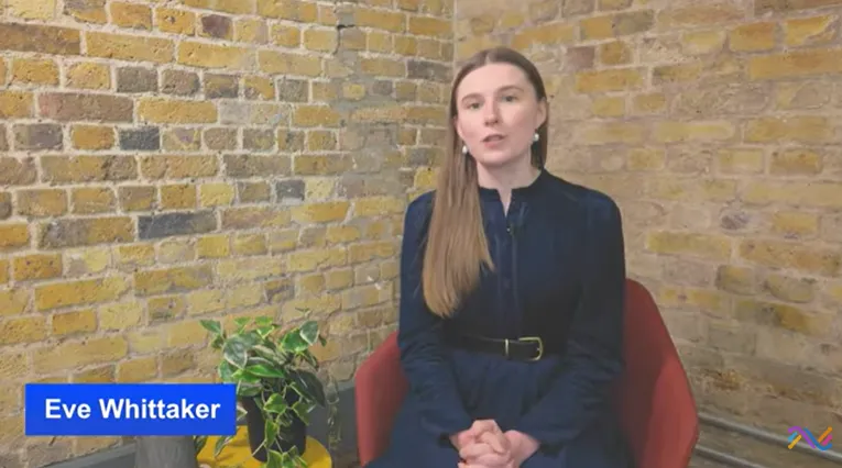 A woman with straight light brown hair, wearing a dark blue dress with a belt, sitting on a red chair in front of a yellow brick wall. A green potted plant is beside her on a yellow table, and a blue name label reads 'Eve Whittaker.'