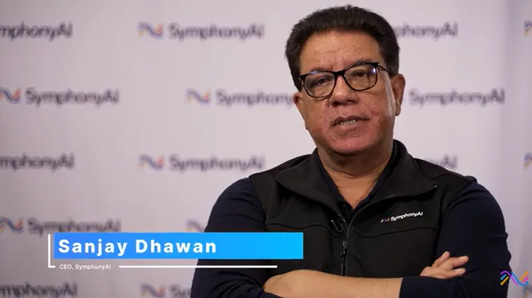 Sanjay Dhawan, CEO of SymphonyAI, wearing glasses and a black sleeveless vest with the SymphonyAI logo, standing with arms crossed in front of a backdrop displaying the SymphonyAI logo repeatedly.