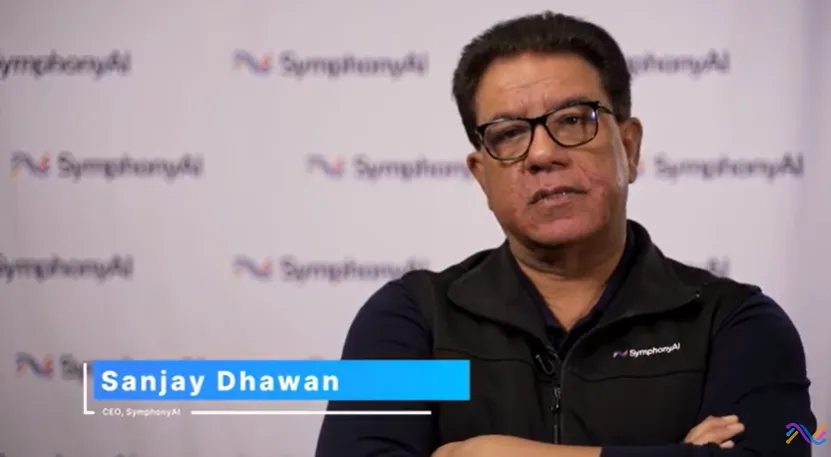 Man with glasses and dark hair, wearing a black SymphonyAI vest over a navy shirt, standing with arms crossed in front of a backdrop featuring the SymphonyAI logo, with a name label reading 'Sanjay Dhawan, CEO, SymphonyAI.'