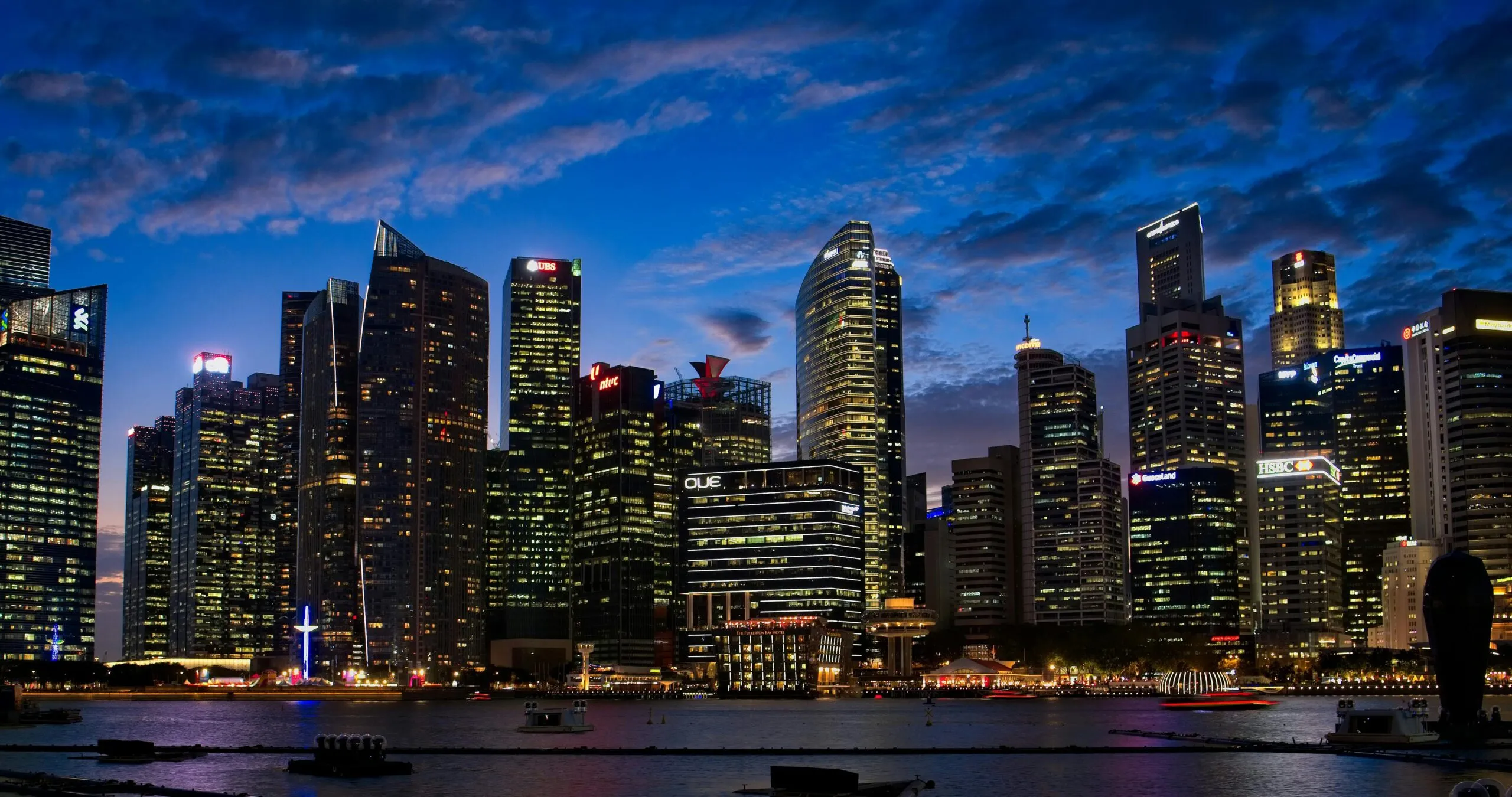 Night view of a modern city skyline with illuminated skyscrapers reflecting on calm water under a partly cloudy blue sky.