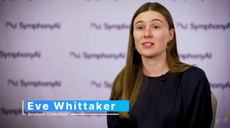 A woman with long brown hair, wearing a dark top, sitting and speaking in front of a light purple backdrop with repeated SymphonyAI logos, with a lower third text overlay identifying her as Eve Whittaker, Solutions Consultant.
