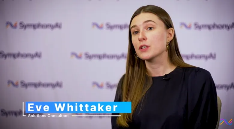 Woman with long brown hair wearing a dark top speaking in front of a backdrop with repeated SymphonyAI logos, identified as Eve Whittaker, Solutions Consultant.