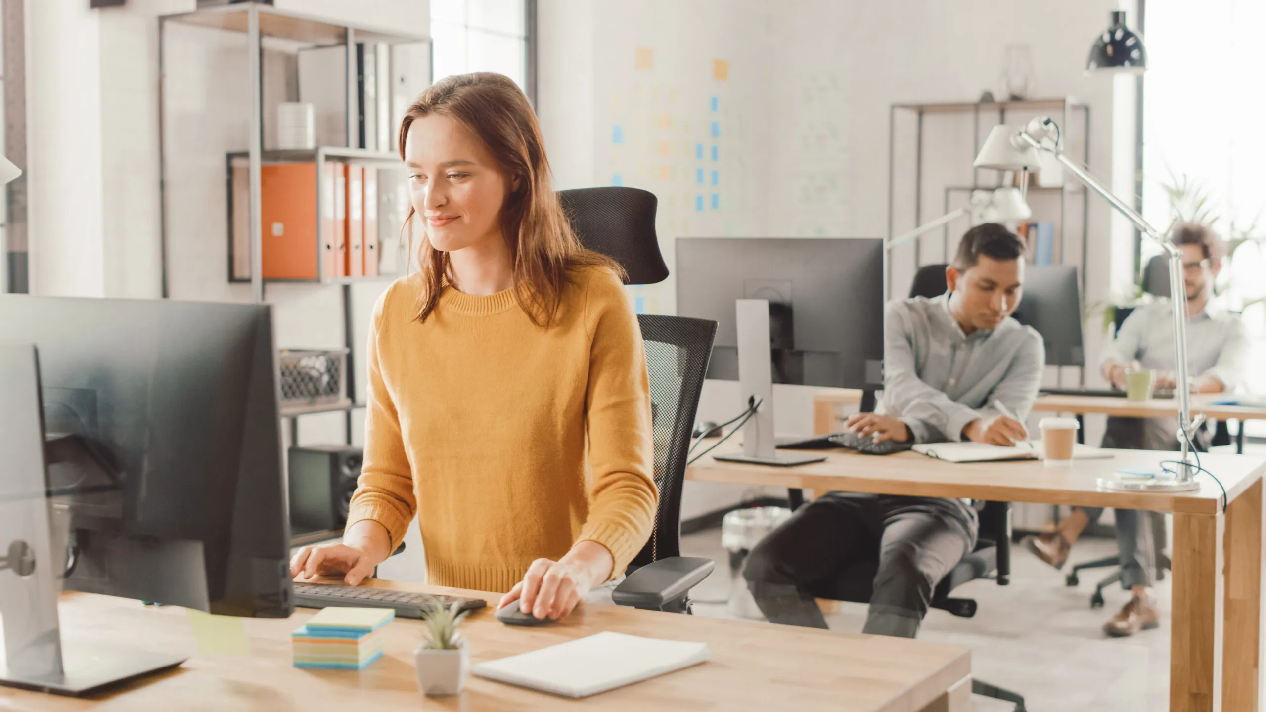 A woman in a yellow sweater sits at a desk using a computer while two men work at desks in the background in a bright, modern office.