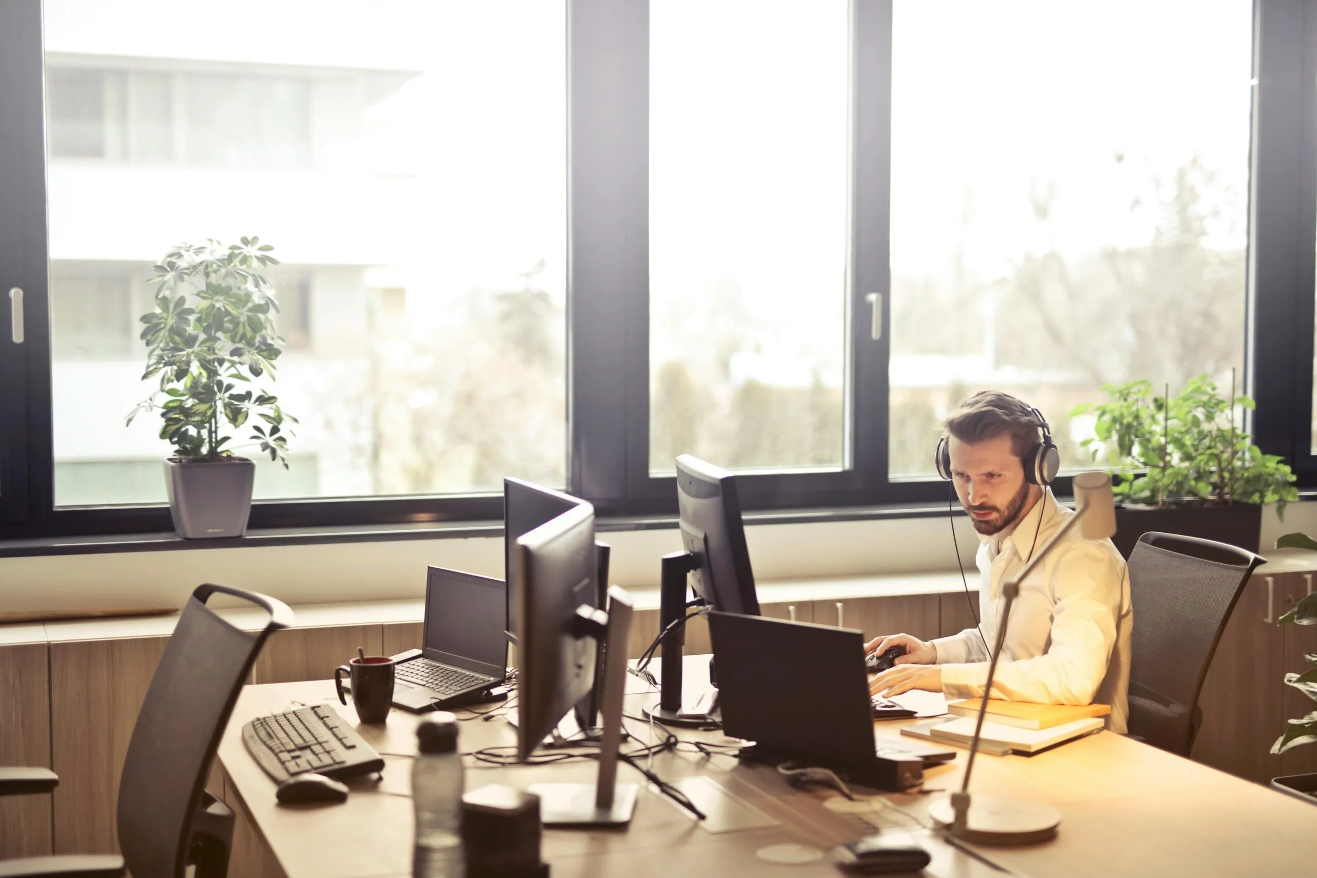 Man wearing headphones and white shirt working on a laptop and computer at a desk in a bright office with large windows, plants, and multiple monitors.