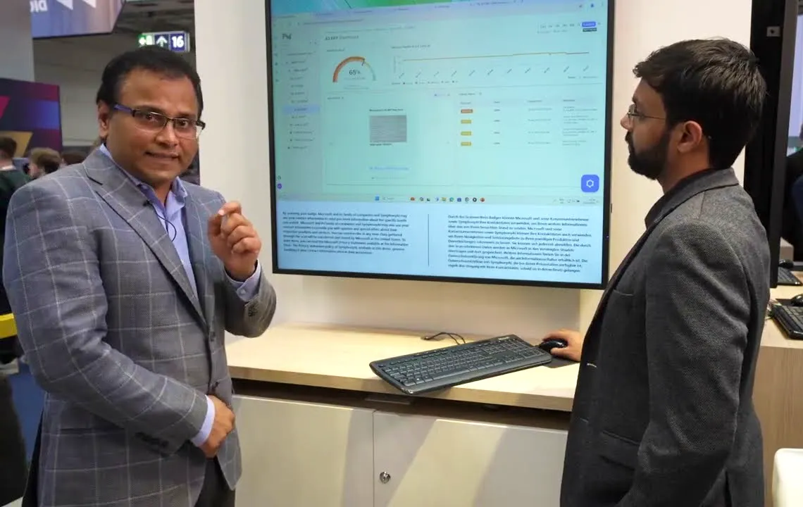 Two men in suits stand in front of a large monitor displaying a dashboard with charts and data tables; one man gestures with his hand while the other uses a computer mouse and keyboard on a desk below the screen.