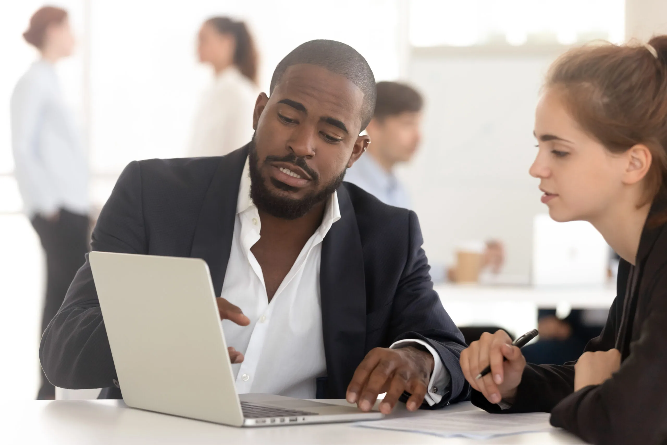 Two colleagues in a modern office discussing work while looking at a laptop, with other people blurred in the background.