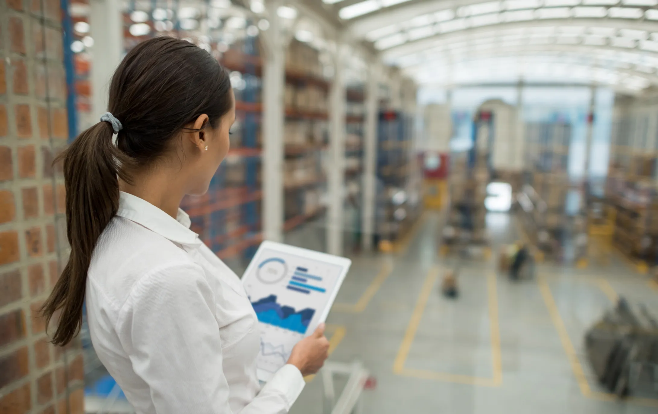 Woman in a white shirt holding a tablet displaying charts and graphs, overlooking a large warehouse with tall shelves and packages.