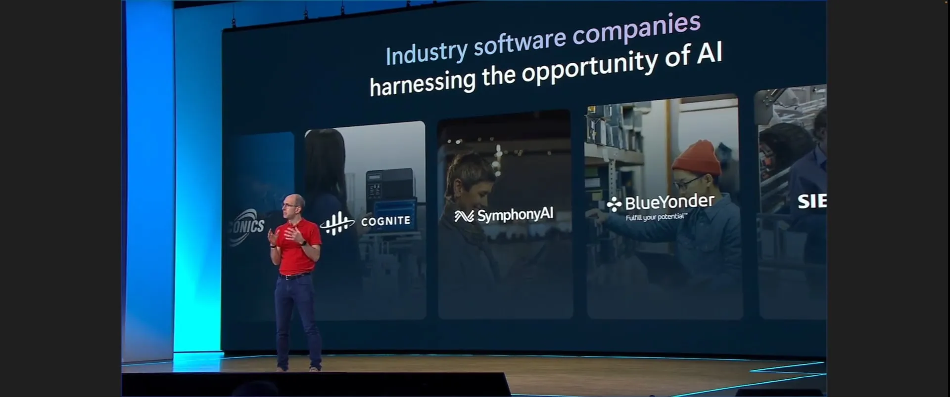 A man in a red shirt and jeans speaking on stage with a large screen behind him displaying the text 'Industry software companies harnessing the opportunity of AI' and logos and images for the companies COGNITE, SymphonyAI, BlueYonder, and Siemens.