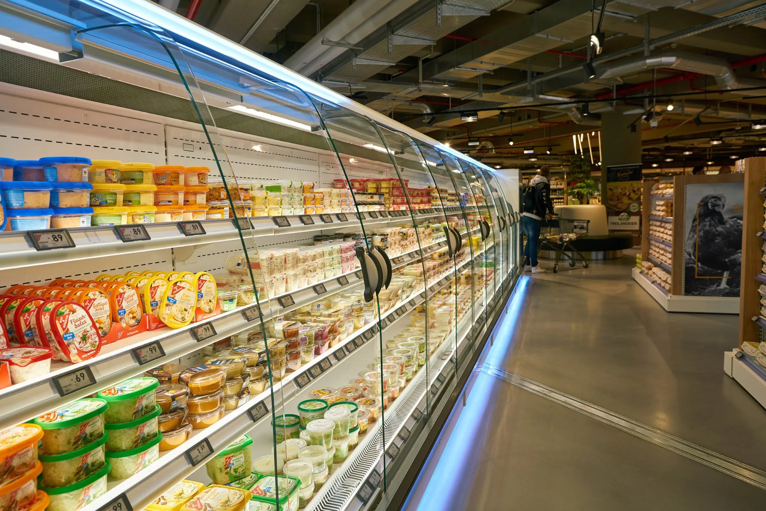 A supermarket aisle with refrigerated glass cases displaying various packaged dairy and salad products on multiple shelves, and a person pushing a shopping cart in the background.