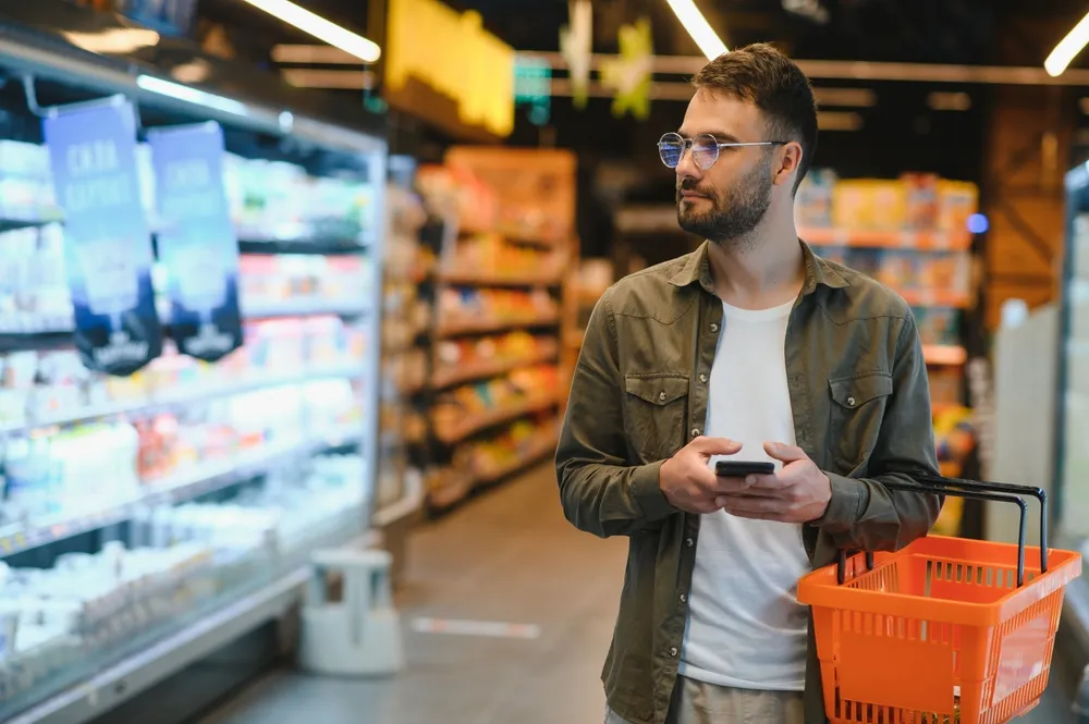 Man with glasses holding a smartphone and an orange shopping basket in a grocery store aisle, looking at refrigerated products on shelves.