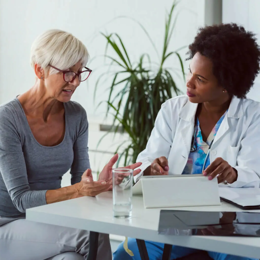 A female doctor in a white coat listens attentively to an older woman with short blonde hair and glasses who is explaining something while seated at a table with a glass of water and a tablet device.