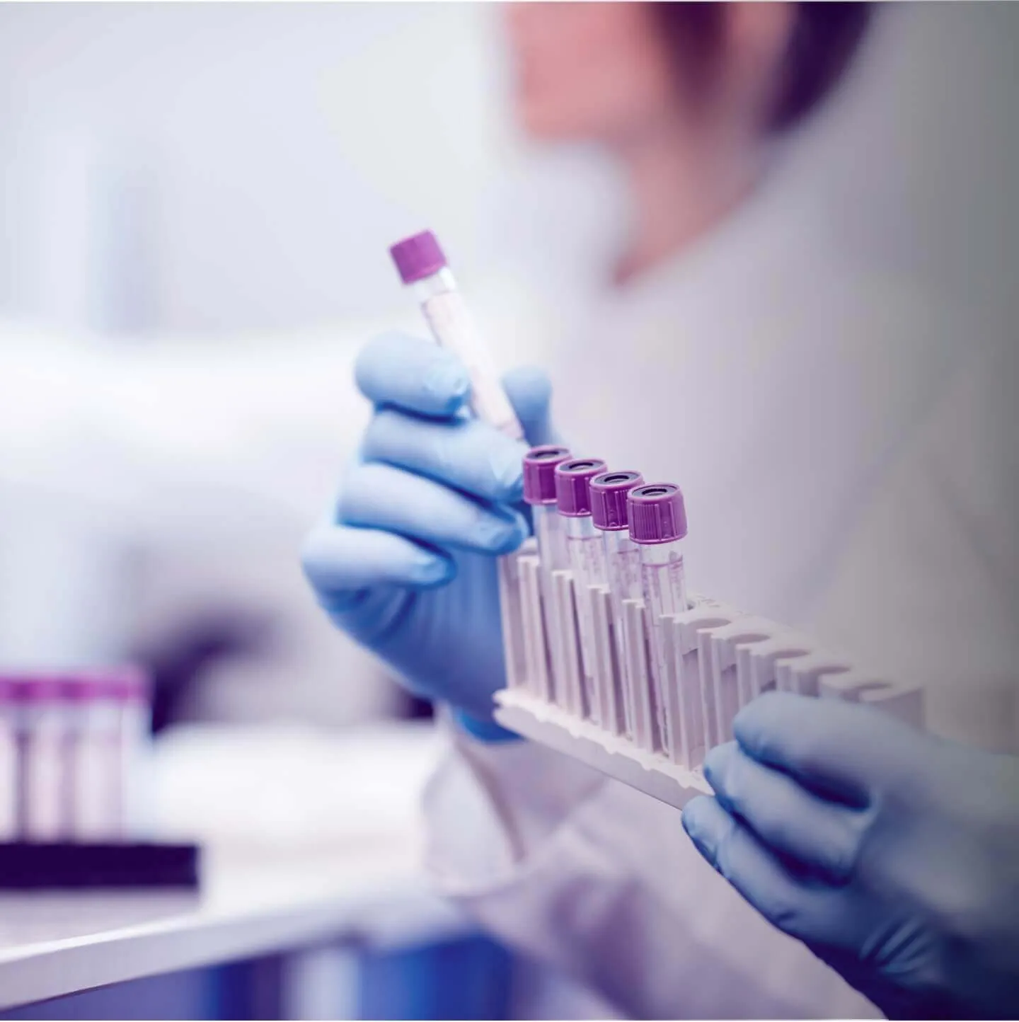Person wearing blue gloves holding a rack with multiple blood sample tubes with purple caps, one tube held separately in the other hand.