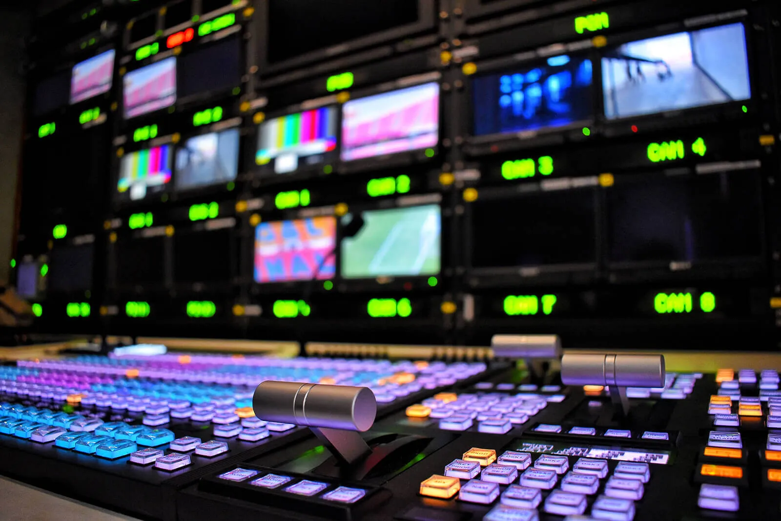 Close-up of a video production control panel with illuminated buttons and two silver levers, multiple screens with colorful test patterns and video feeds in the background.